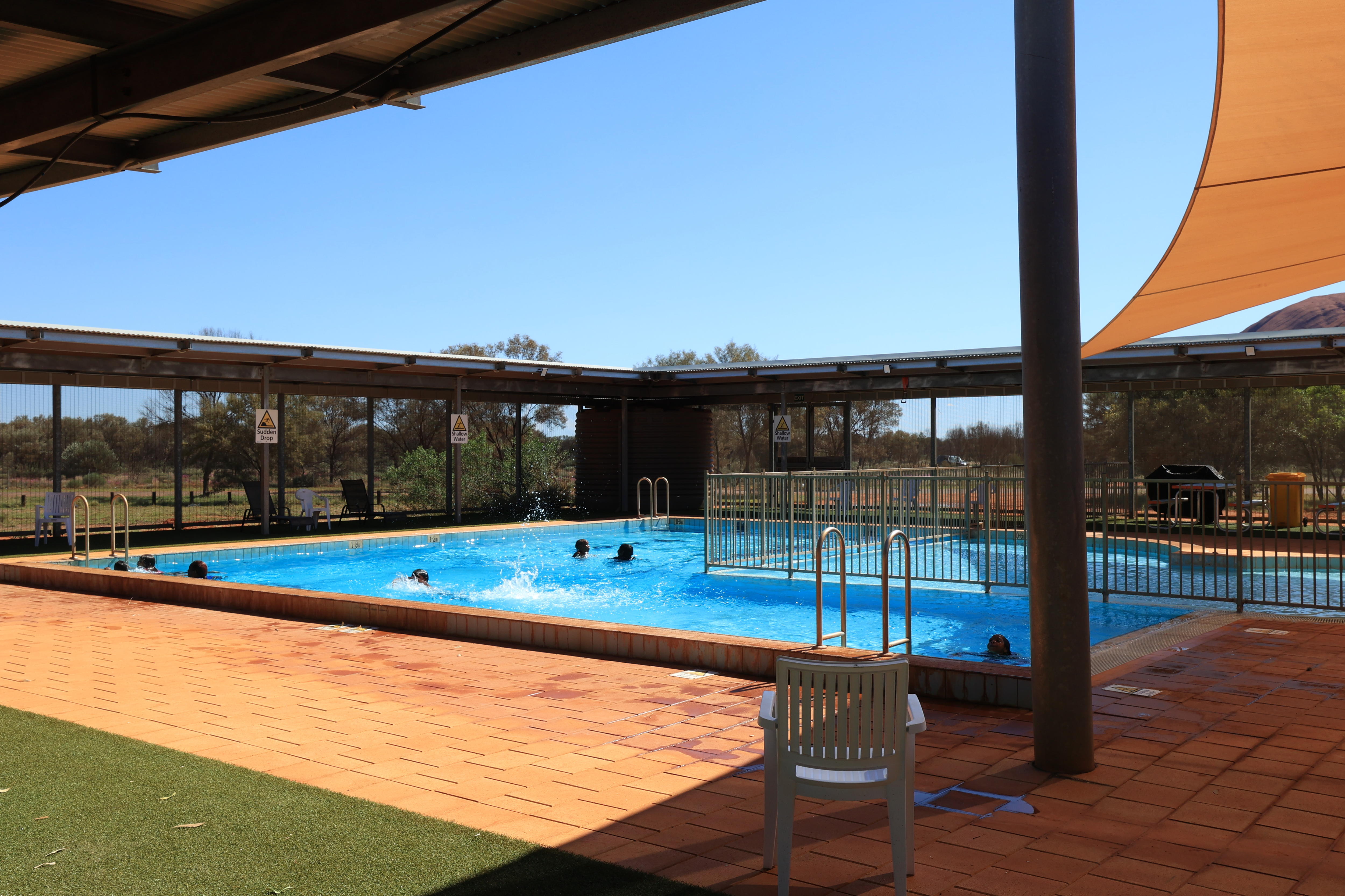 Children splash around in a public pool, there's a shade cloth overhead and a rocky hill can be seen in the background.