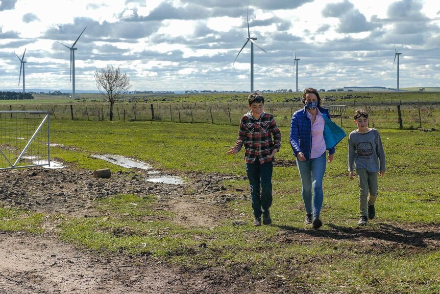 A woman in blue jeans and purple jacket walks through a muddy paddock with her two sons at her side, wind turbines in background
