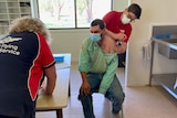 a man sitting on a chair getting  a vaccine, as a RFDS staff member works at a table.