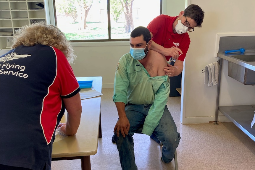 a man sitting on a chair getting  a vaccine, as a RFDS staff member works at a table.