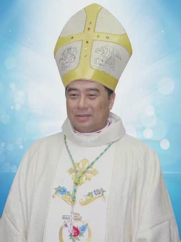 A man wearing a light-coloured robe with embroidery and a crucifix around his neck poses for a portrait