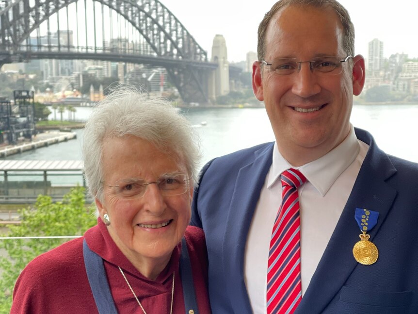 An older, bespectacled woman and a younger, dark-haired man in a suit smile, the Sydney Harbour Bridge behind them.
