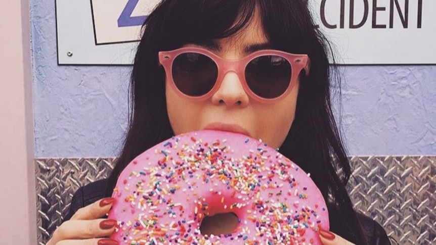 A woman bites into a large, pink iced donut.