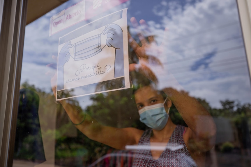 A woman wearing a face mask holds up a sign to a window. The sign shows two hands holding a mask.
