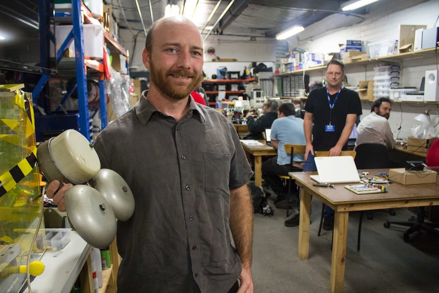 A man holds an electronic bell in a room filled with technology and tools, with other men in the background