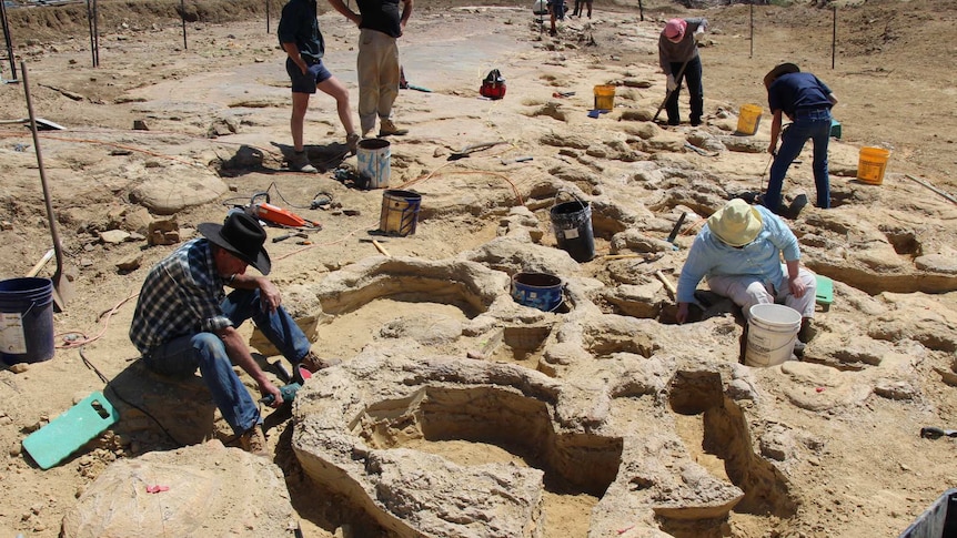 People with buckets and tools sit in the dirt, digging around deep divots in the dusty stone.