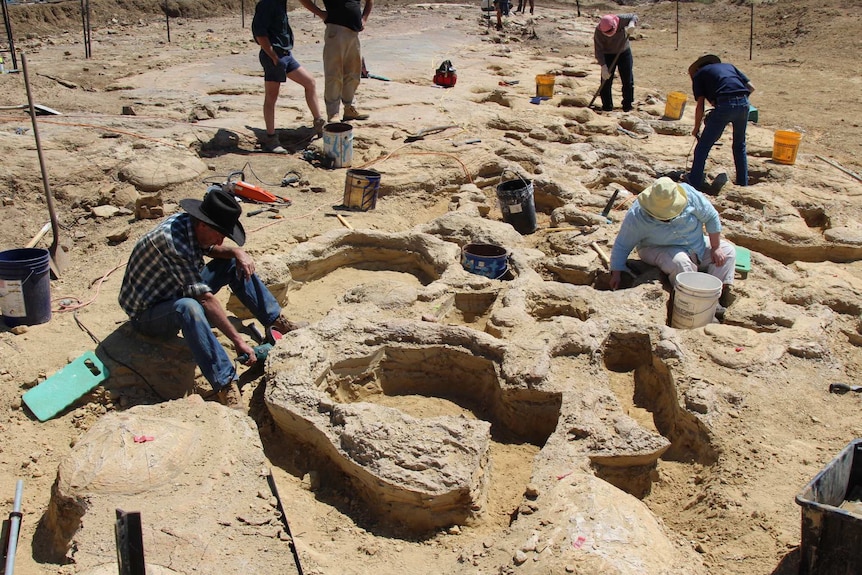 People with buckets and tools sit in the dirt, digging around deep divots in the dusty stone.