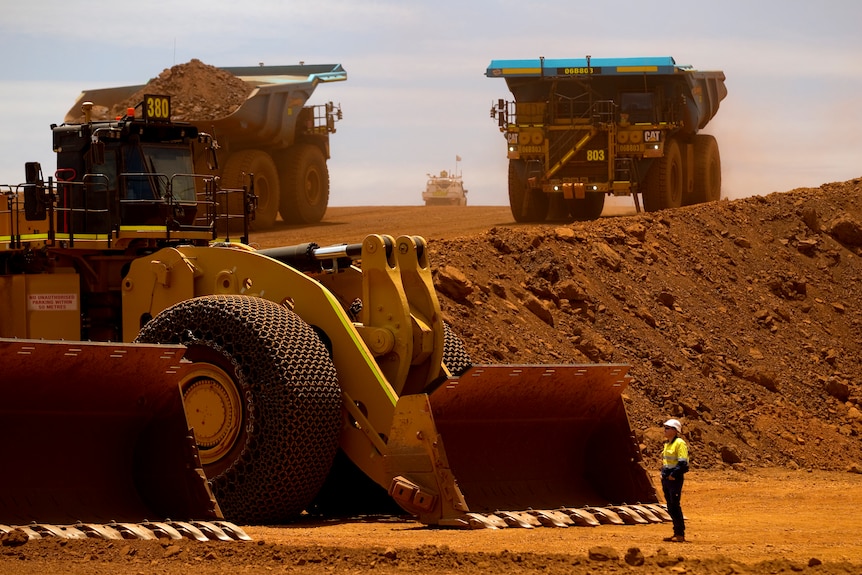 A man in bright yellow is dwarfed by three very large trucks in red dirt