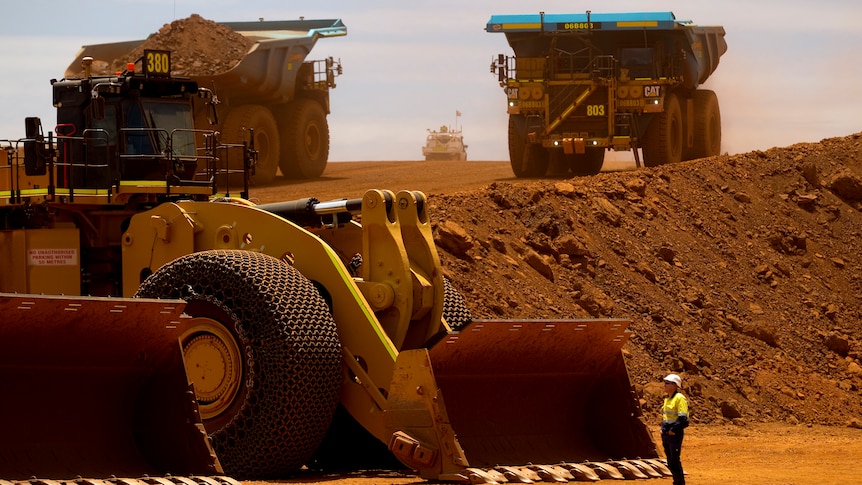 A man in bright yellow is dwarfed by three very large trucks in red dirt