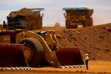 A man in bright yellow is dwarfed by three very large trucks in red dirt