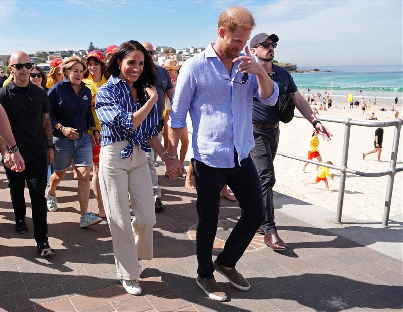 Harry and Meghan walk along Bondi Beach with surf life savers behind them.