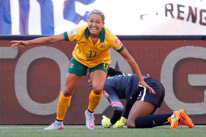A Matildas striker smiles as she picks herself up after scoring a goal, with the goalkeeper still on the ground.