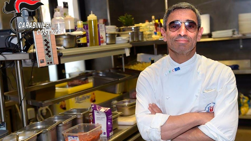 A man in a chef's coat and sunglasses stands in a kitchen with his arms folded. 