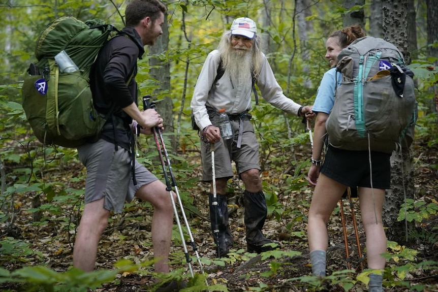 A young man and women speak with an older man with long great hair on a lush, green hiking trail 