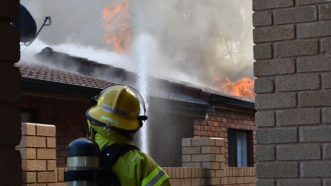 A firefighter standing next door to a house of fire. Large amounts of smoke and flames can be seen.