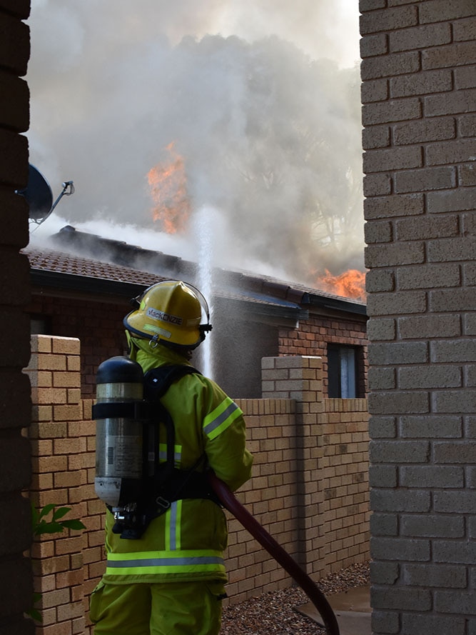 A firefighter standing next door to a house of fire. Large amounts of smoke and flames can be seen.