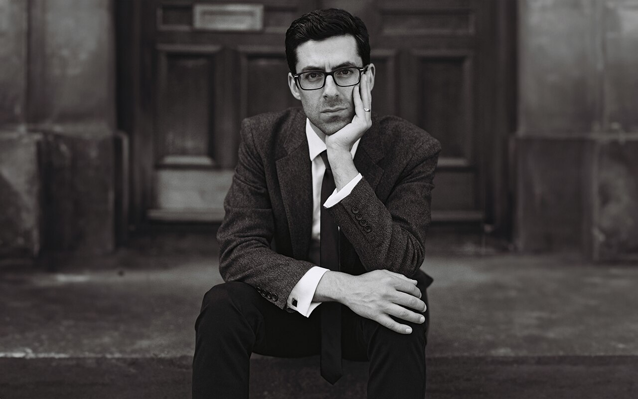 Gabriel Latchin sits on a step and stares directly into the camera. He is in a suit and is leaning his head on his hand.