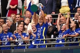 Sam Kerr roars in triumph as she lifts a big trophy above her head and teammates applaud. 