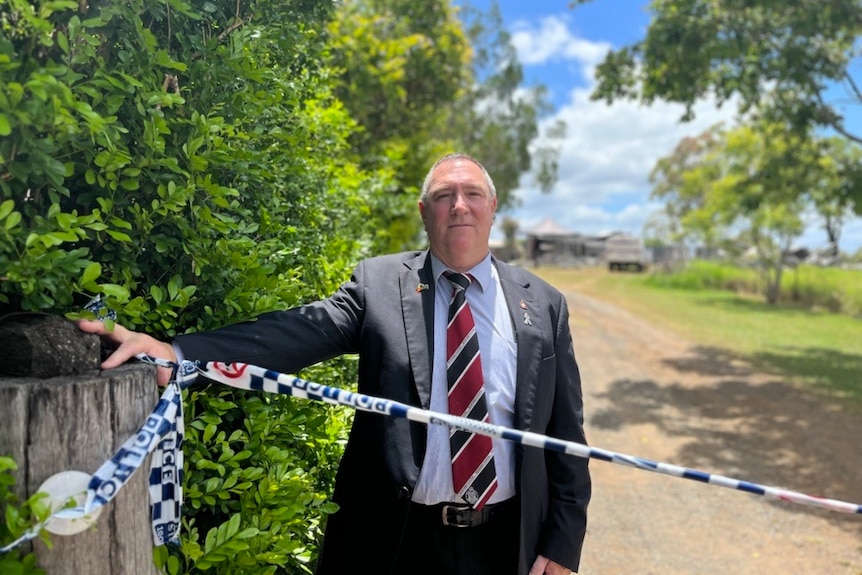 A man in a suit looks at the camera, police tape in front of him, leaning against a post with a property behind