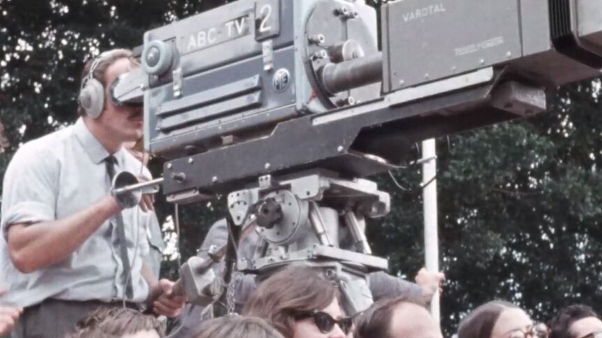 A man operates a large, bulk television camera on a platform elevated above a crowd.