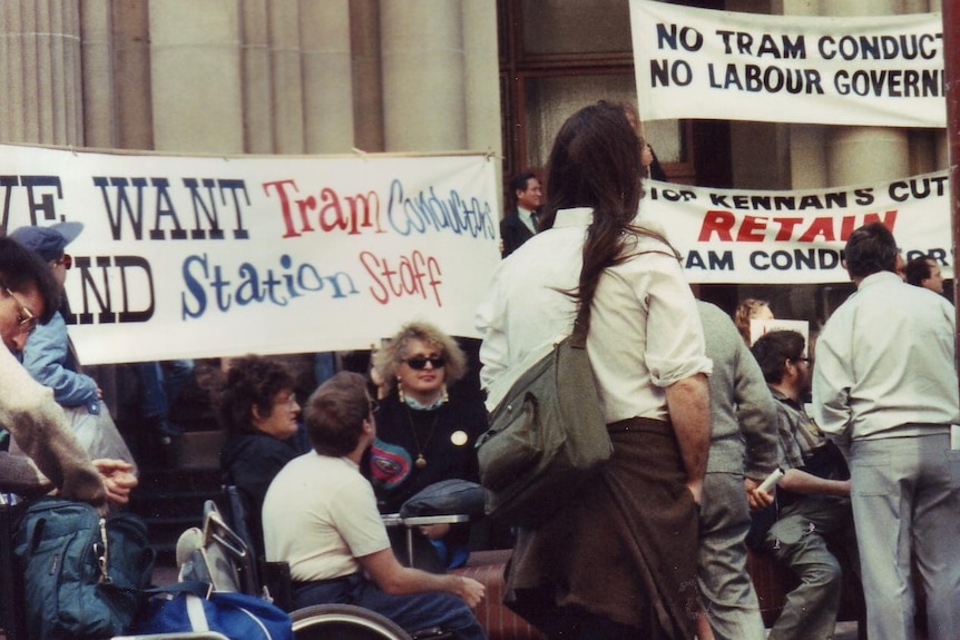 Protesters in wheelchairs gather outside a formal building bearing signs calling for better accessibility on public transport.