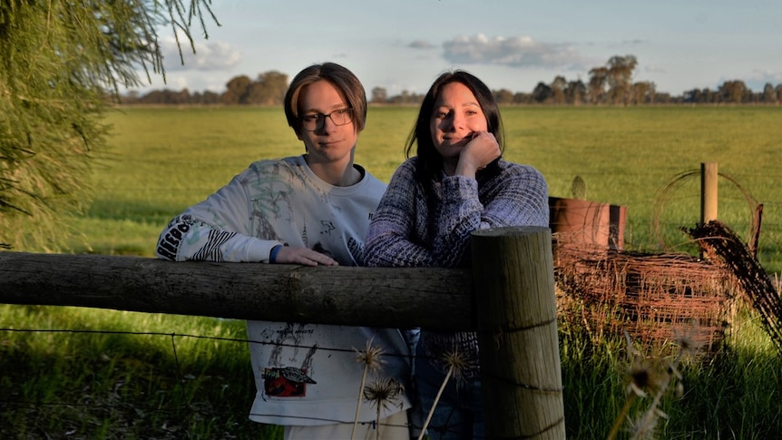 A smiling woman and a pensive teenage boy rest their arms on a farm fence, blue sky, green paddock behind.