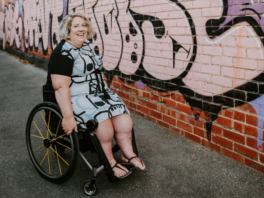 A woman in a wheelchair smiles at the camera, she's in front of a brick wall with prominent graffiti.