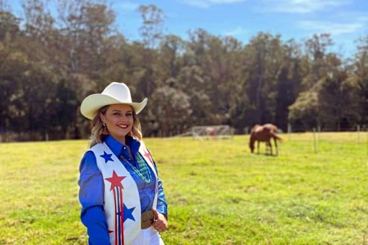 Lady wearing cowgirl hat and blue top stands in field with a horse in the background. 