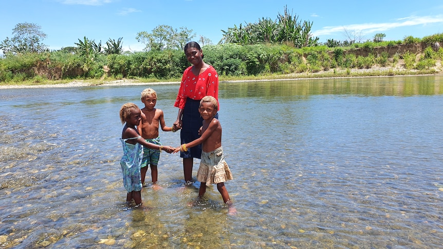 A teenage girl and three young children, hold hands while standing in a body of water.