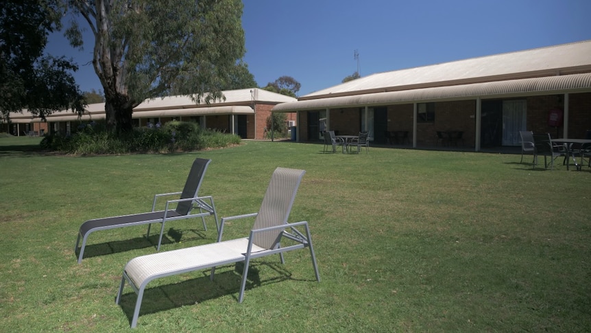 Two empty lounge chairs on a green lawn with apartments behind them. No people are in sight.