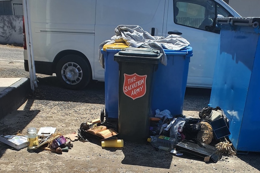 Rubbish lies around wheelie bins with a 'Salvation Army' sticker, white van behind