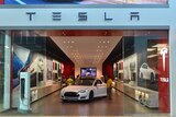Store glass doors part to show a white Tesla sedan with its doors open as people look into it.