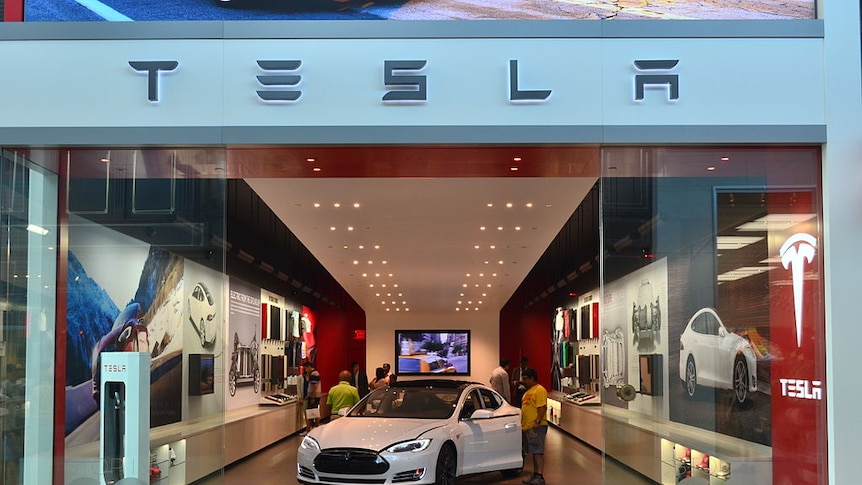 Store glass doors part to show a white Tesla sedan with its doors open as people look into it.