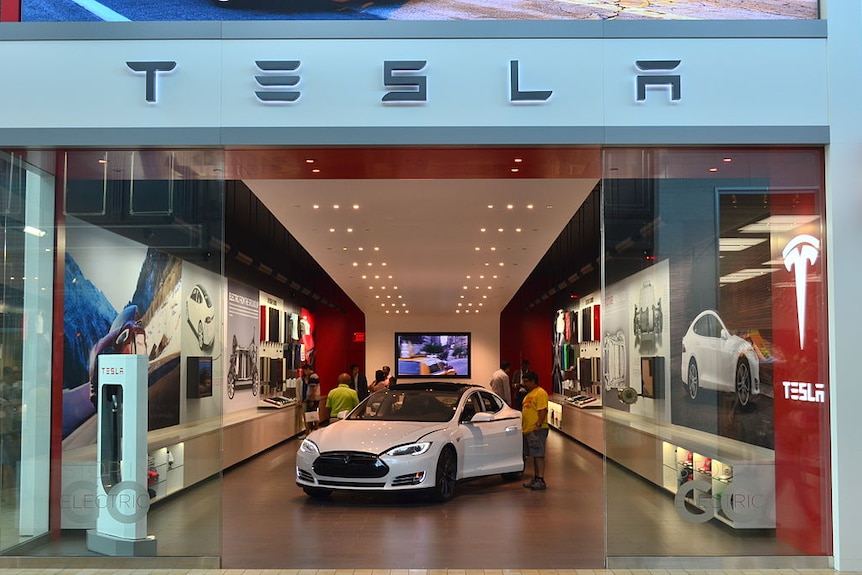 Store glass doors part to show a white Tesla sedan with its doors open as people look into it.