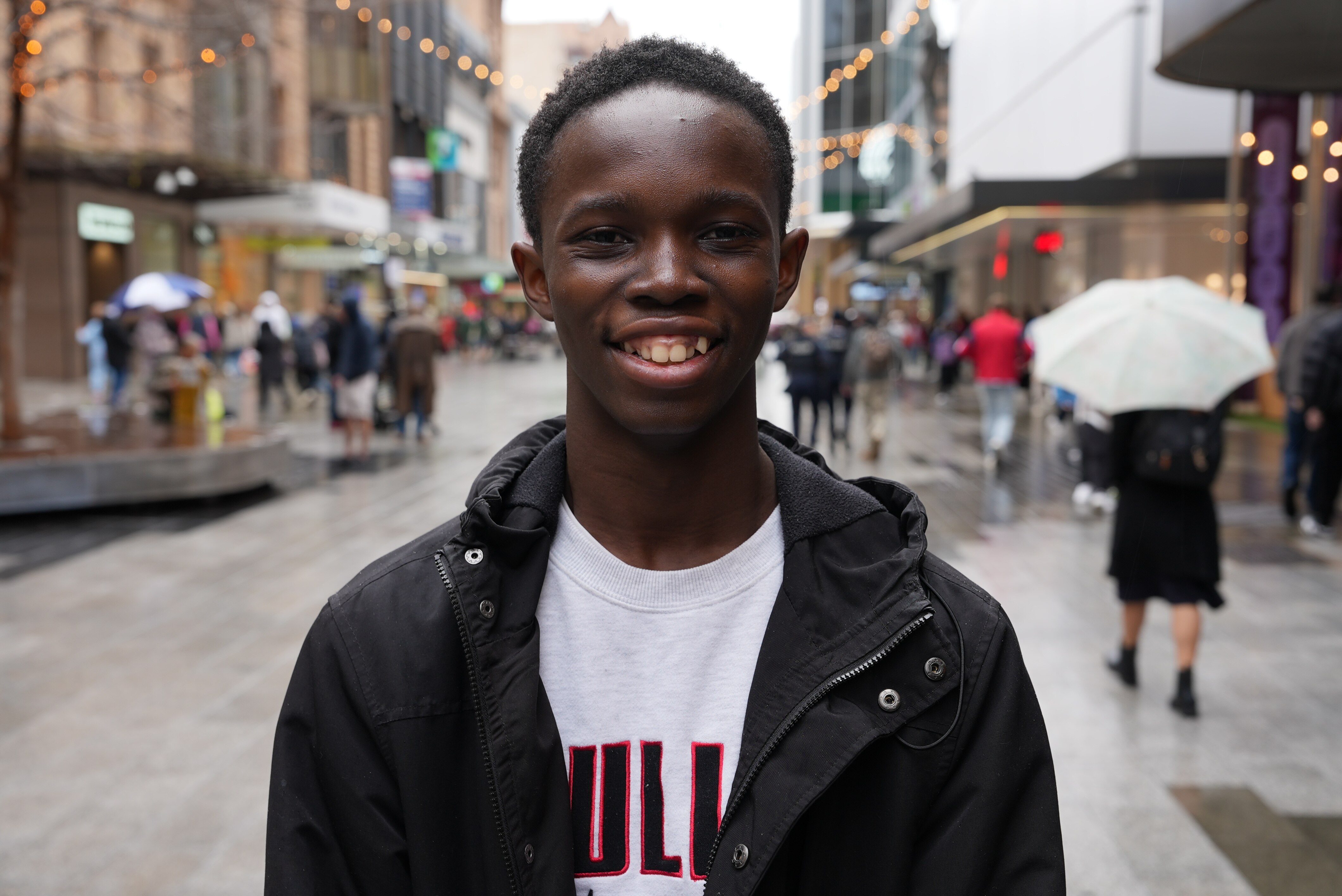 A young smiling young man stands in an outdoor shopping mall where it has recently been raining.