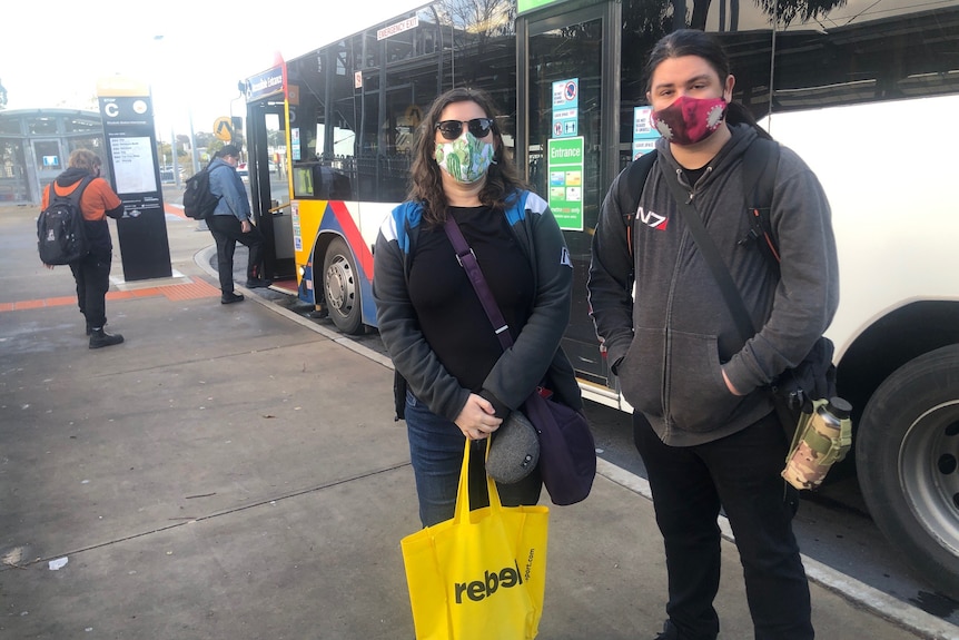 A woman and a man wearing face masks stand in front of a bus at a bus stop