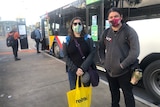 A woman and a man wearing face masks stand in front of a bus at a bus stop