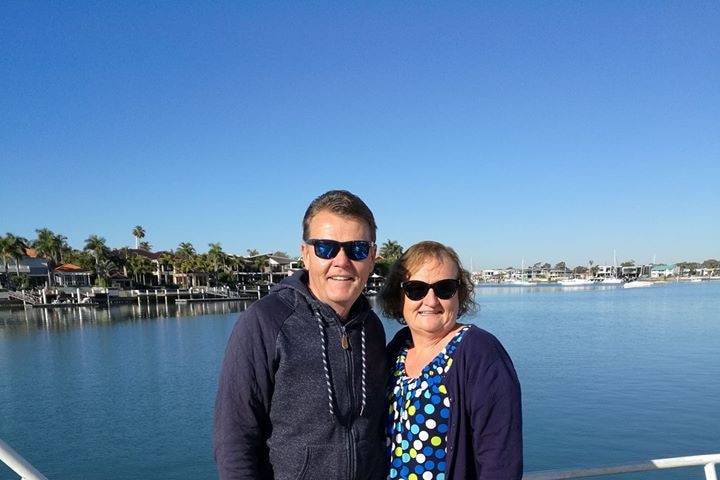 Paul and Coralie Williamson stand aboard a boat with sunglasses on hugging each other and smiling.