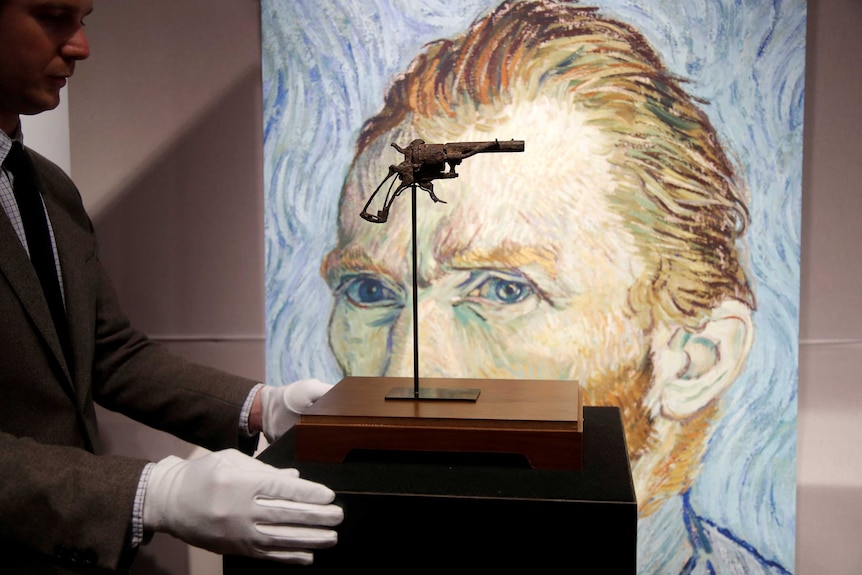 A man in white gloves positions a rusty revolver in front of a self-portrait of the artist Vincent Van Gogh