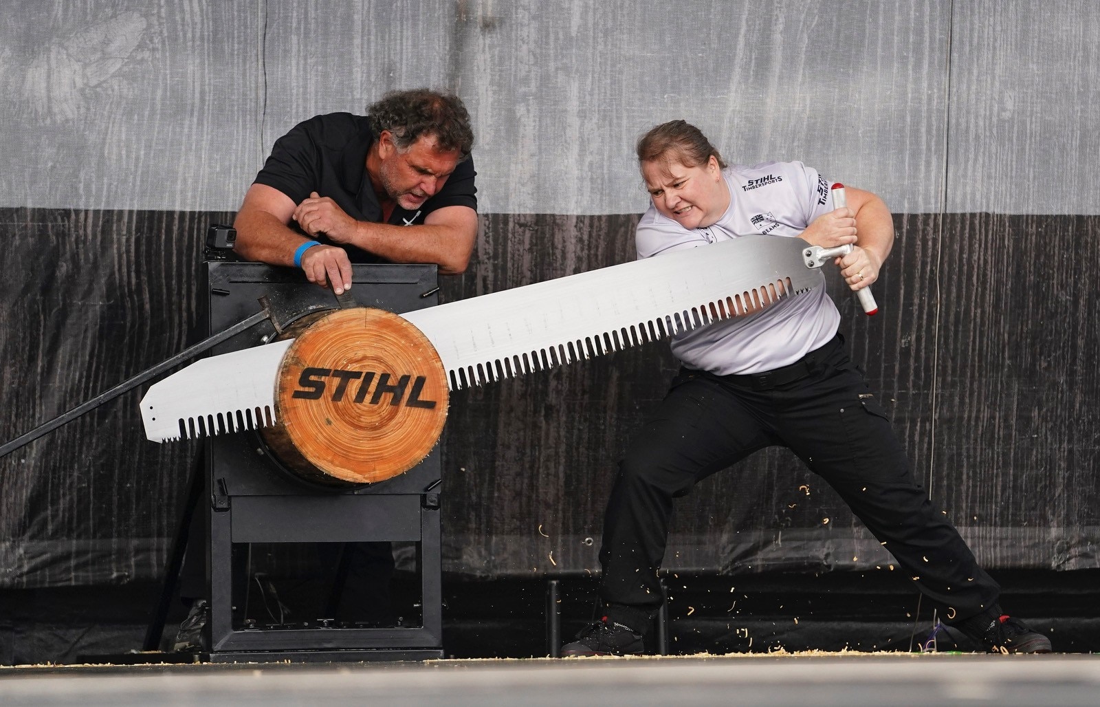 mulher vestindo camiseta cinza e calças vazias fica na extremidade de uma grande serra, com um homem ao lado dela apoiando-a 