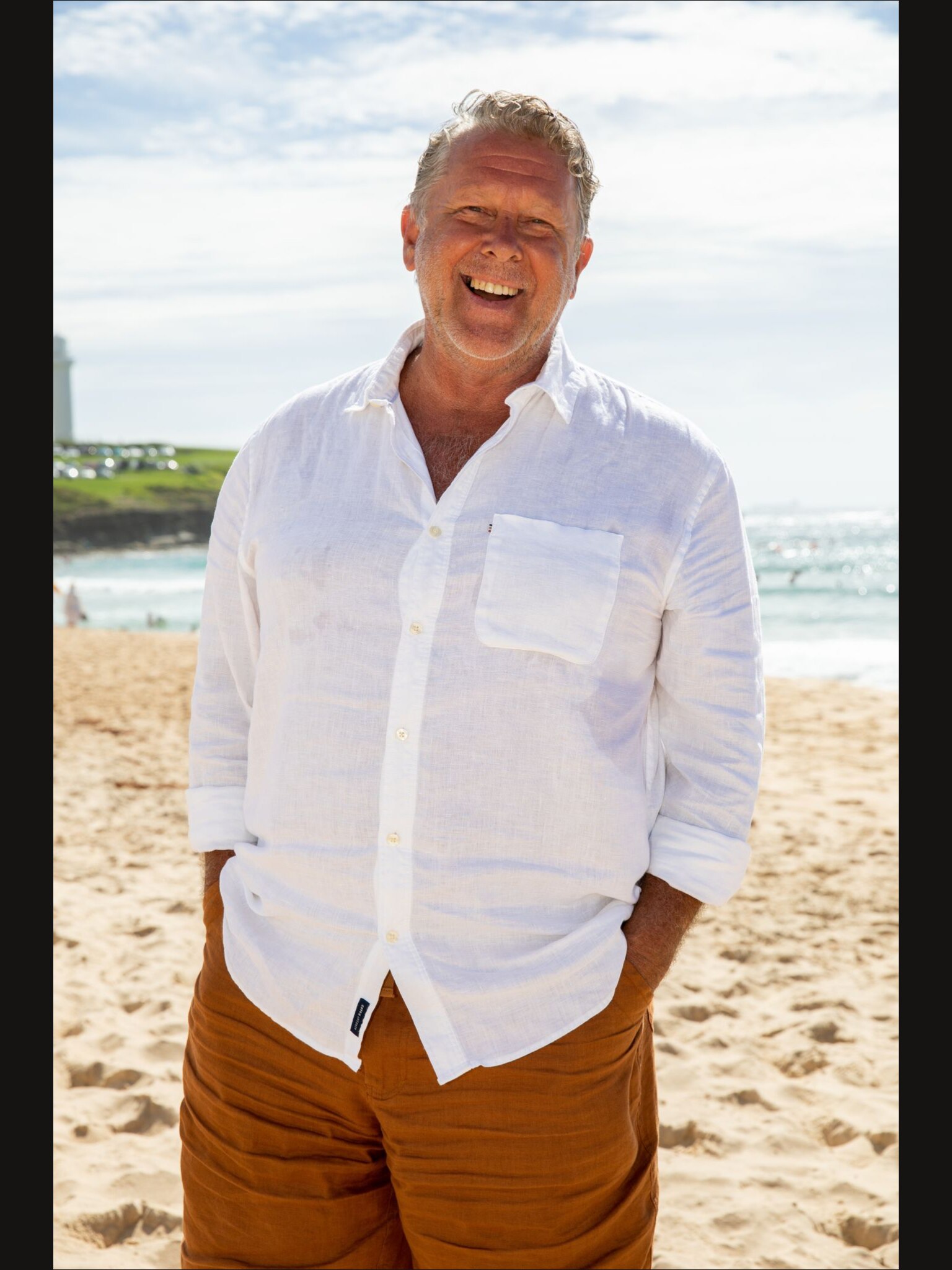 Brad Farmer wears a white button up shirt and camel coloured shorts and smiles with his hands in his pockets on a beach.