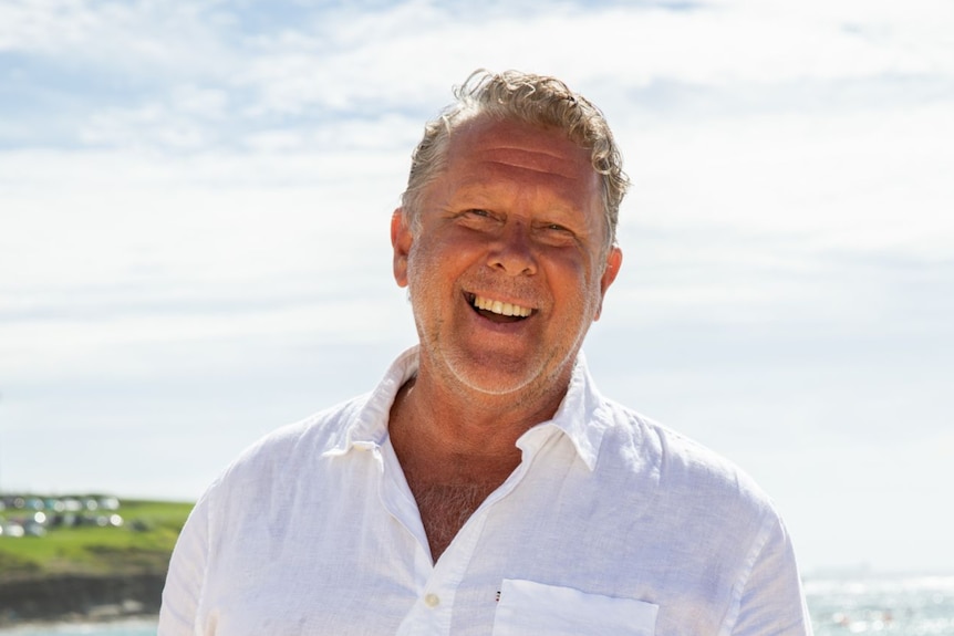 Brad Farmer viste una camisa blanca con botones y pantalones cortos color camel y sonríe con las manos en los bolsillos en la playa.