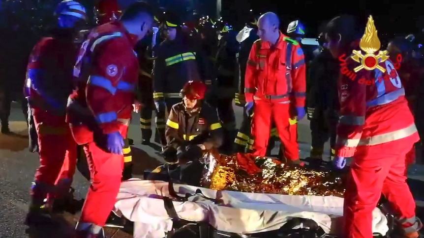 Four people in bright uniforms marked with a red cross look down at as another tends to a person on a stretcher