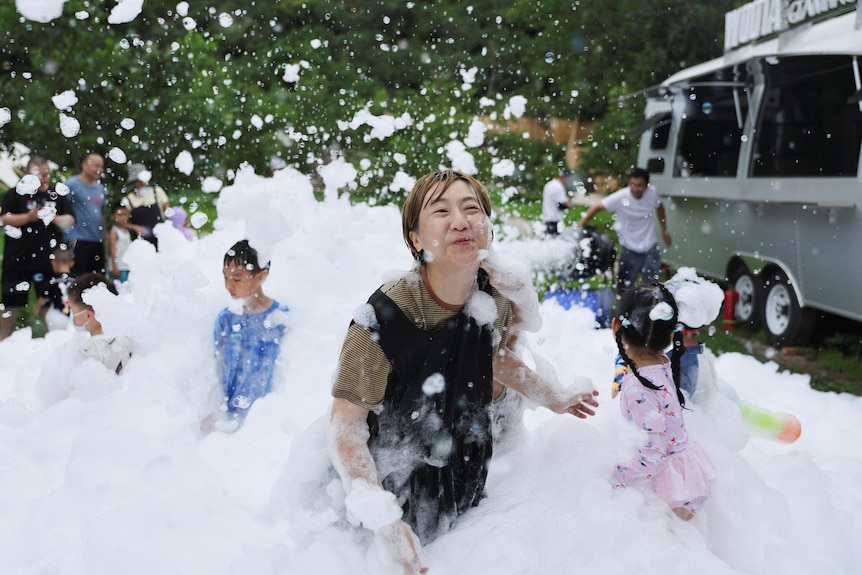 A woman blows bubbles and in the background young children blow bubbles. 