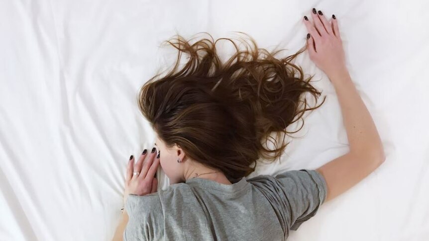 Women with long brown hair, weraing a white t-shirt sleeping on a large bed with white sheets 