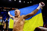 Boxer Oleksandr Usyk holds a Ukrainian flag of blue and yellow behind him as he smiles in victory after a world title fight.