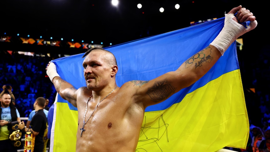 Boxer Oleksandr Usyk holds a Ukrainian flag of blue and yellow behind him as he smiles in victory after a world title fight.