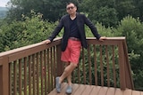 Yang Hengjun wears red shorts, a dark t shirt and black blazer as he stands on a deck with trees and cloudy skies in background.