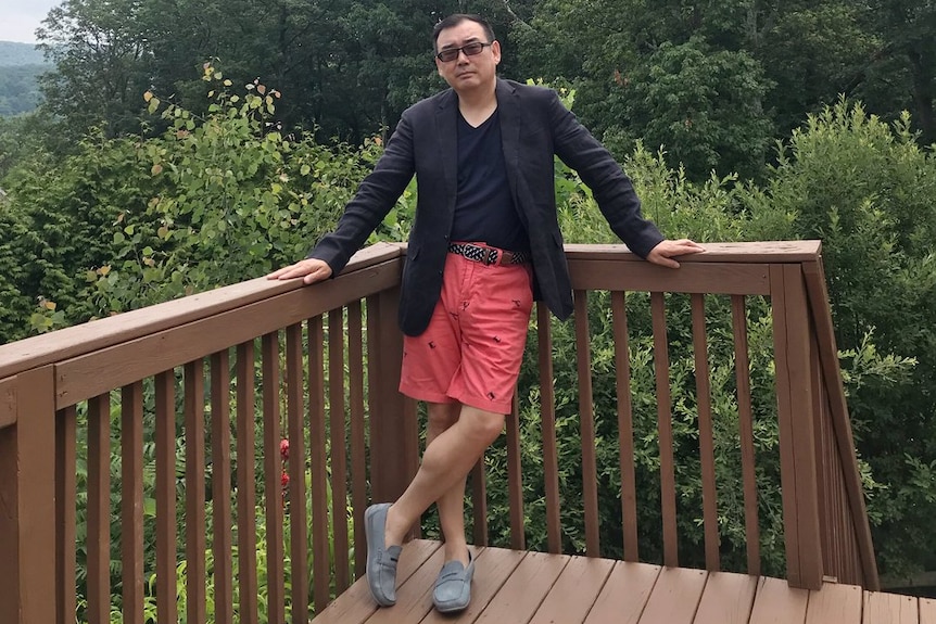 Yang Hengjun wears red shorts, a dark t shirt and black blazer as he stands on a deck with trees and cloudy skies in background.