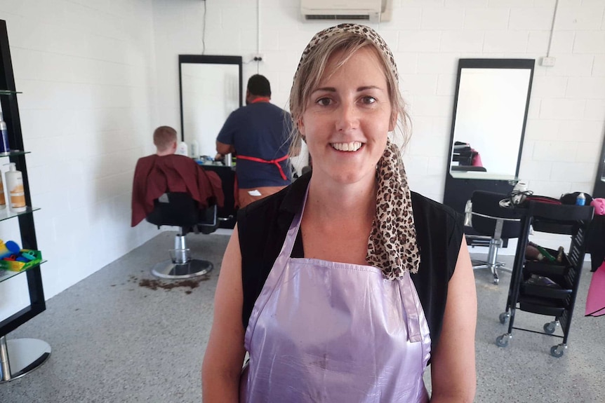A woman wearing a leopard print headscarf and purple apron in a salon smiles.